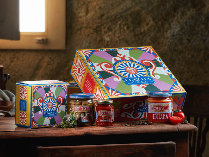 Two Cunzata boxes in carretto colours on a wooden table, with jars of conserves and cherry tomatoes