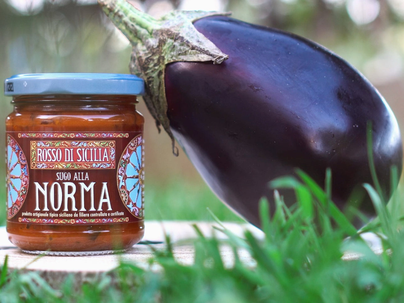 A jar of Sugo alla Norma on a round of wood in the grass, beside a whole aubergine