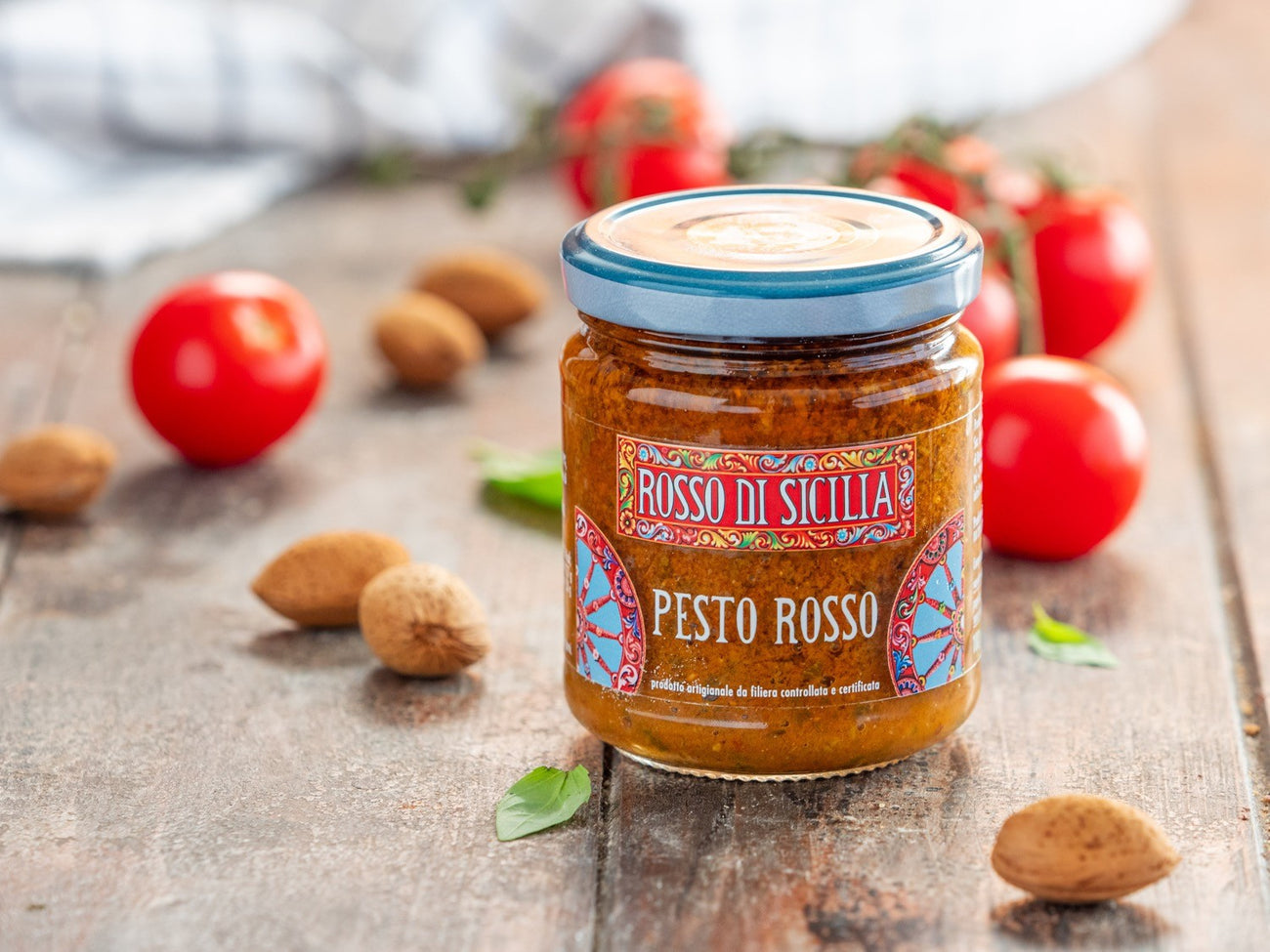 A jar of Pesto Rosso on a wooden table, with almonds, cherry tomatoes and basil leaves