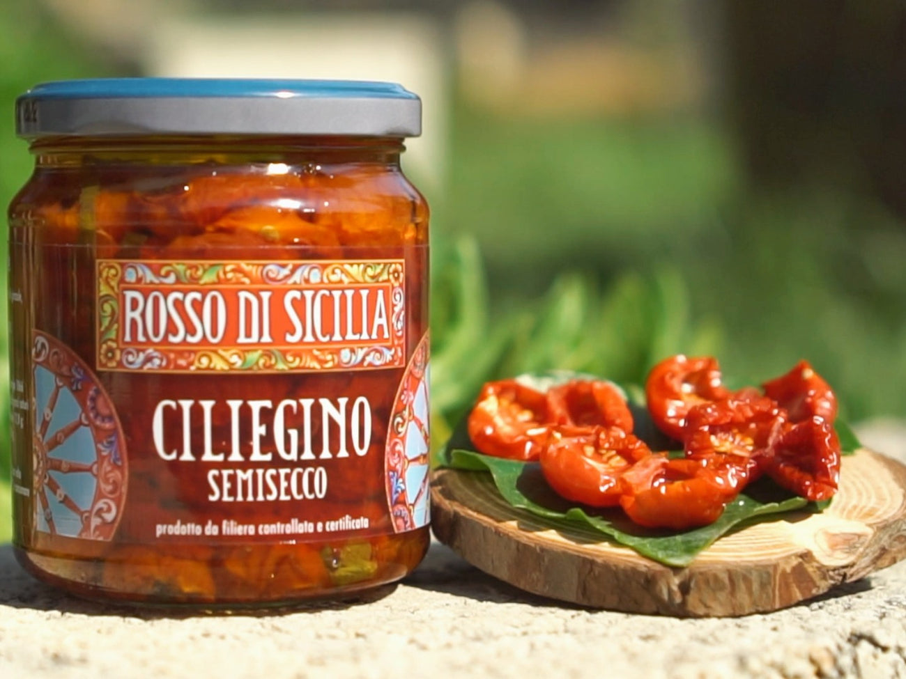 A jar of semi-dried cherry tomatoes on a stone ledge, beside a few on a leaf and a round wooden board