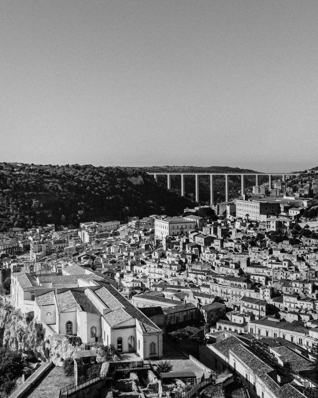 Black-and-white view over the rooftops of Modica with the Guerrieri viaduct above the town