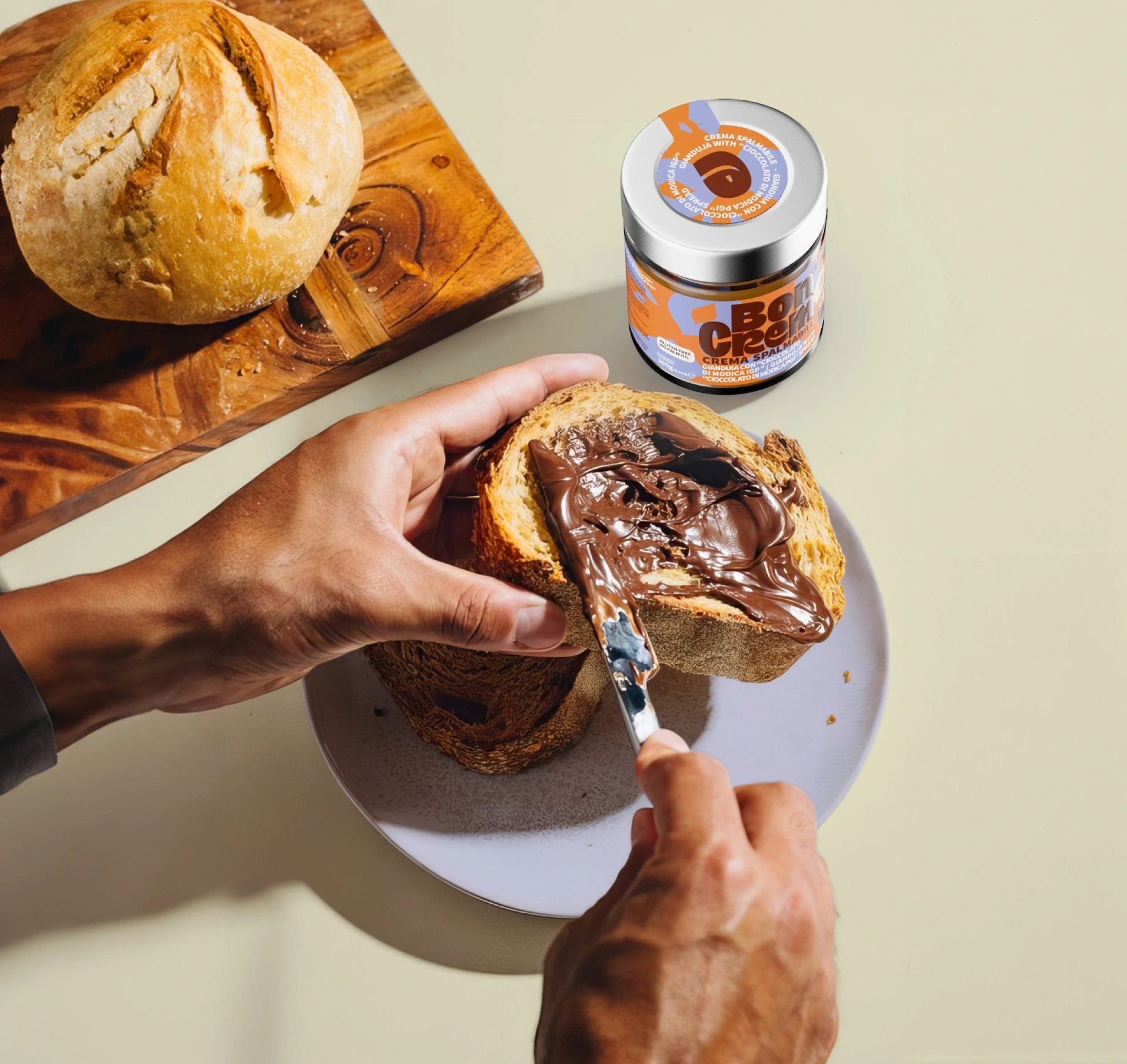 Hands spreading BonCrem hazelnut and cocoa cream on a slice of Sicilian bread, with the jar and a wooden board alongside.