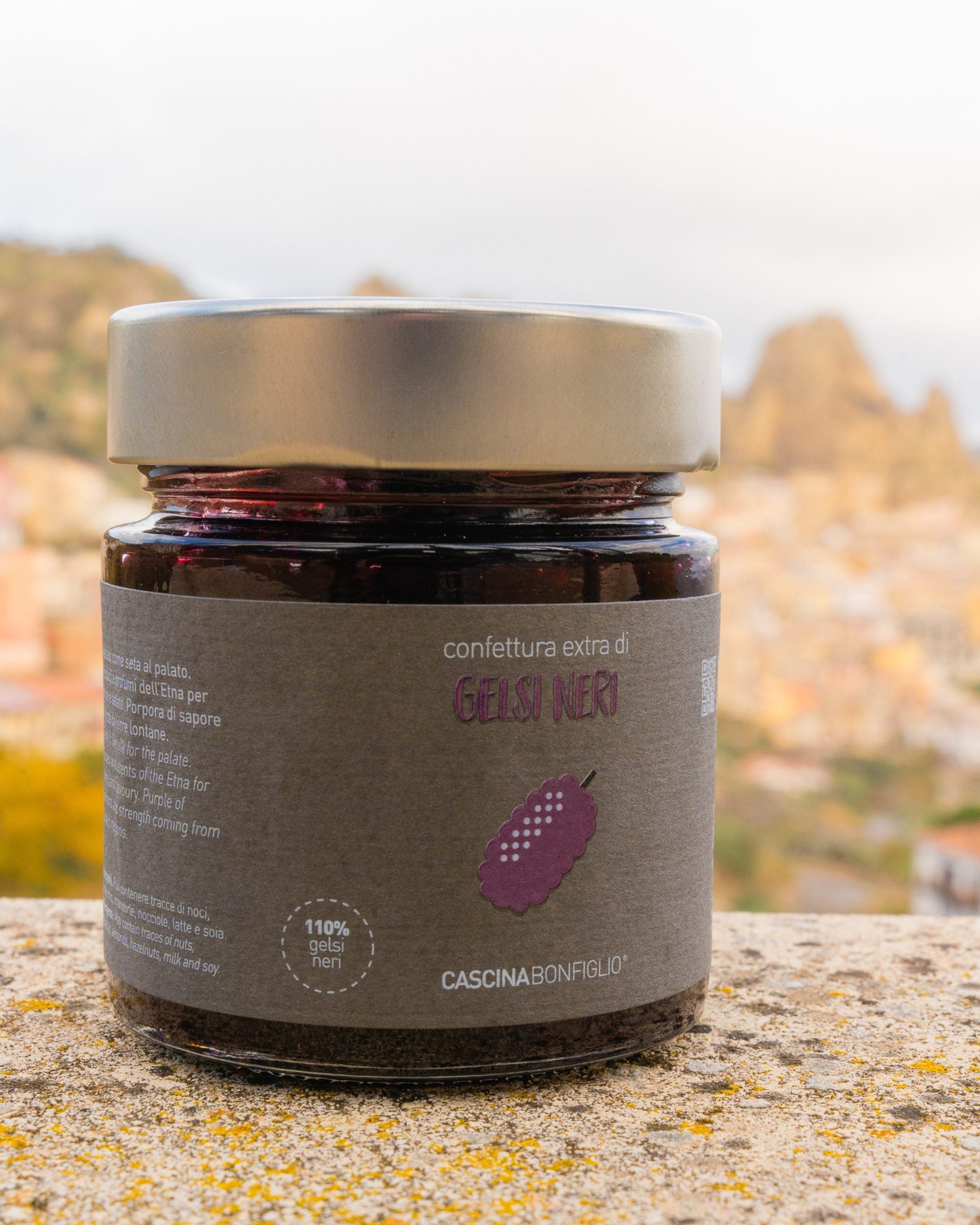 A jar of Cascina Bonfiglio black mulberry preserve on a stone wall, with a Sicilian hill town behind it