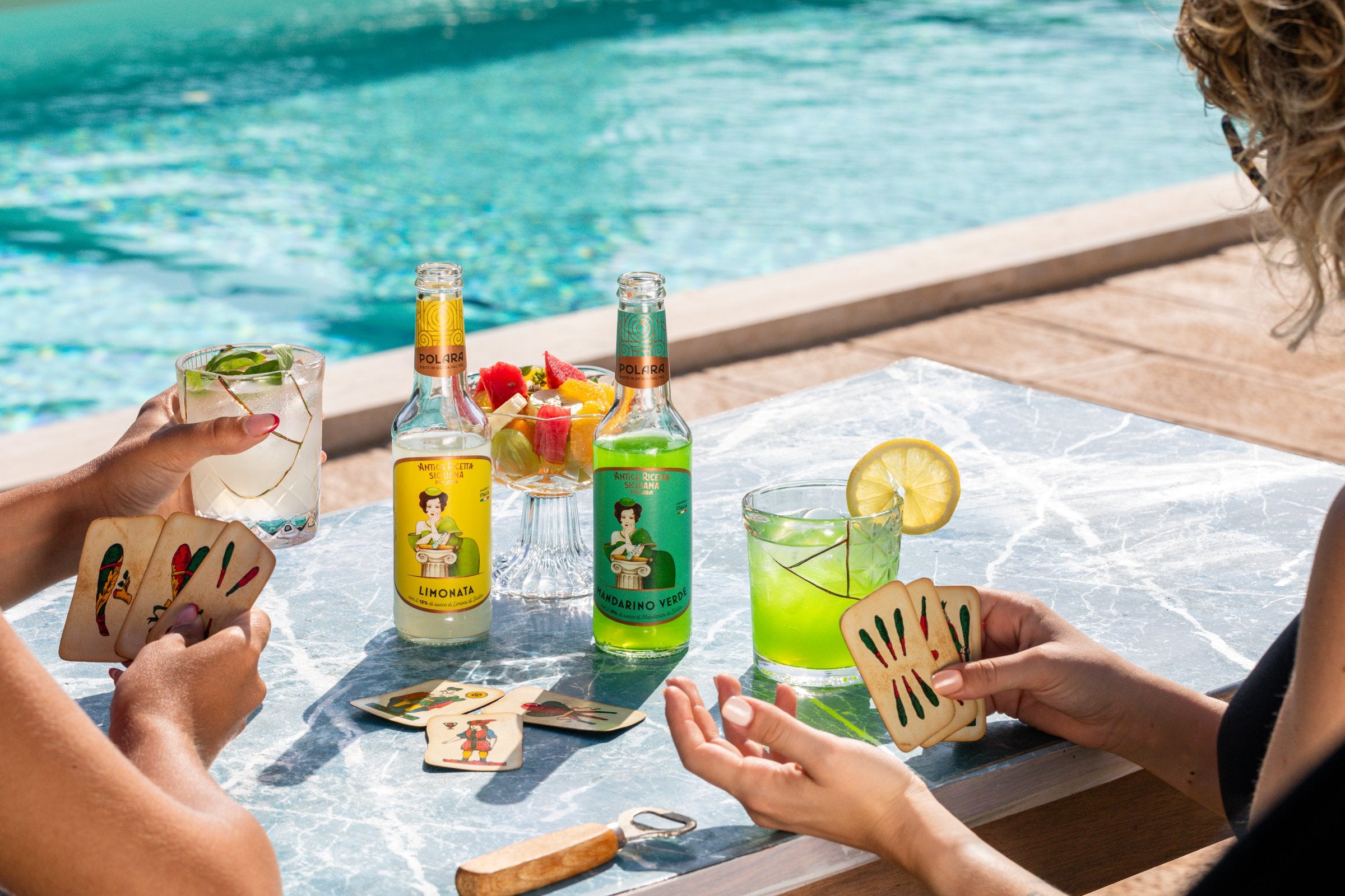 Hands holding Sicilian playing cards beside bottles of Polara Limonata and Mandarino Verde