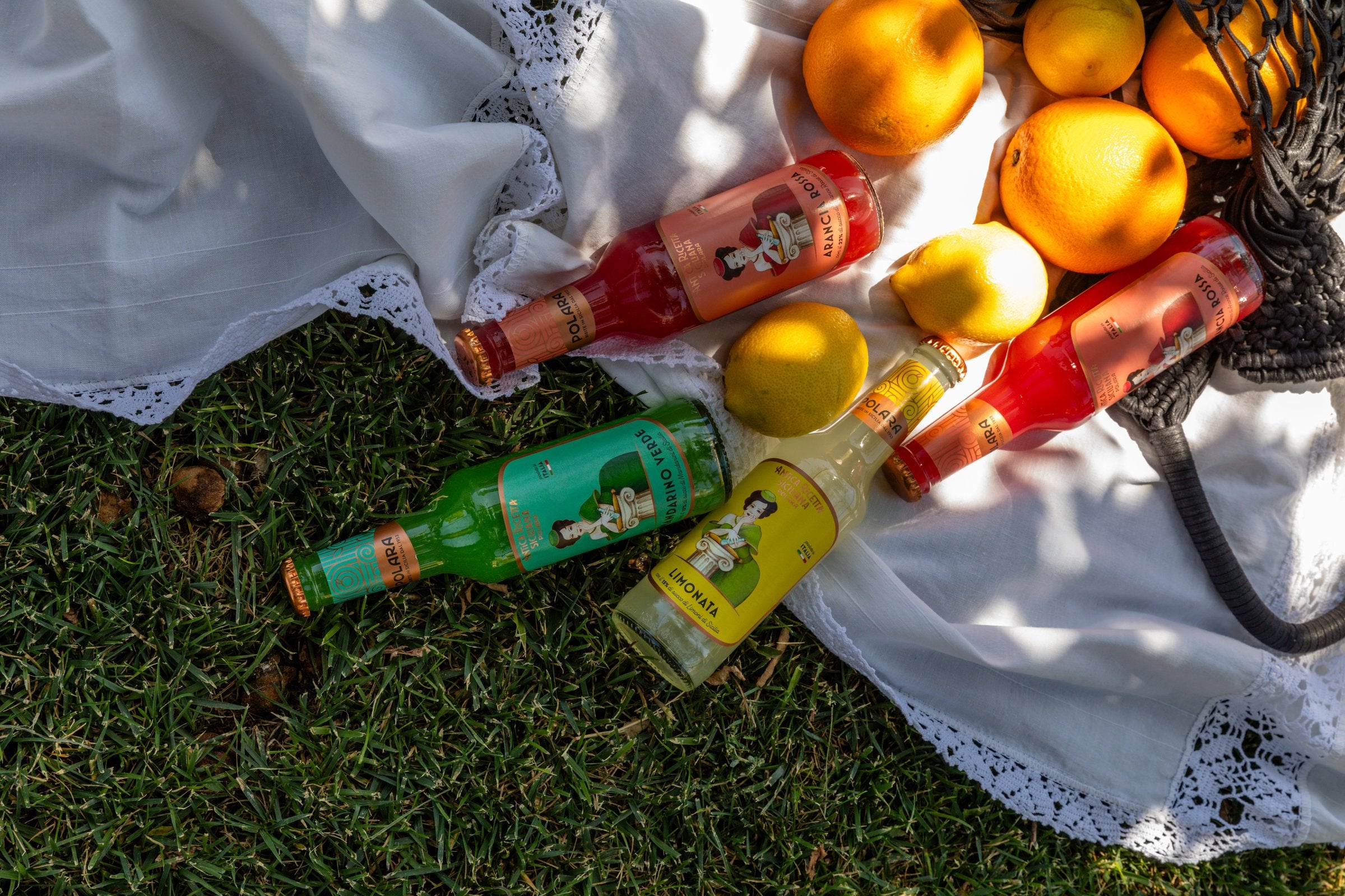 Bottles of Polara lying on the grass among oranges, lemons and a net bag at a picnic