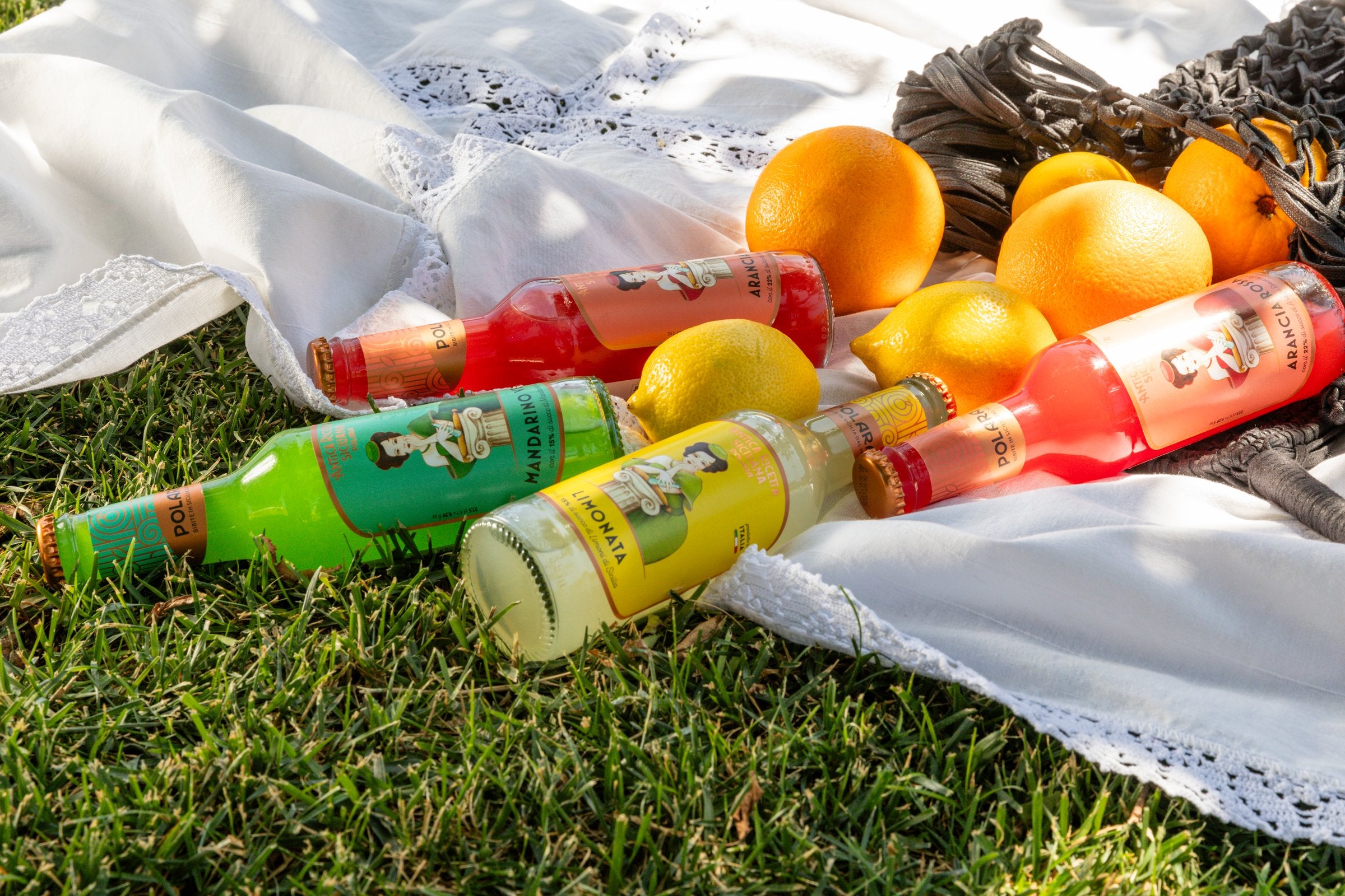 Bottles of Polara lying on the grass at a picnic, with oranges, lemons and a net bag