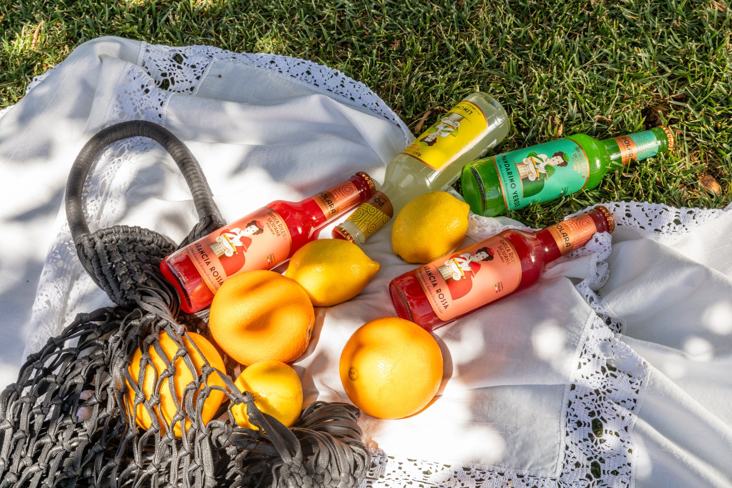 A picnic on the grass with bottles of Polara, oranges and lemons in a net