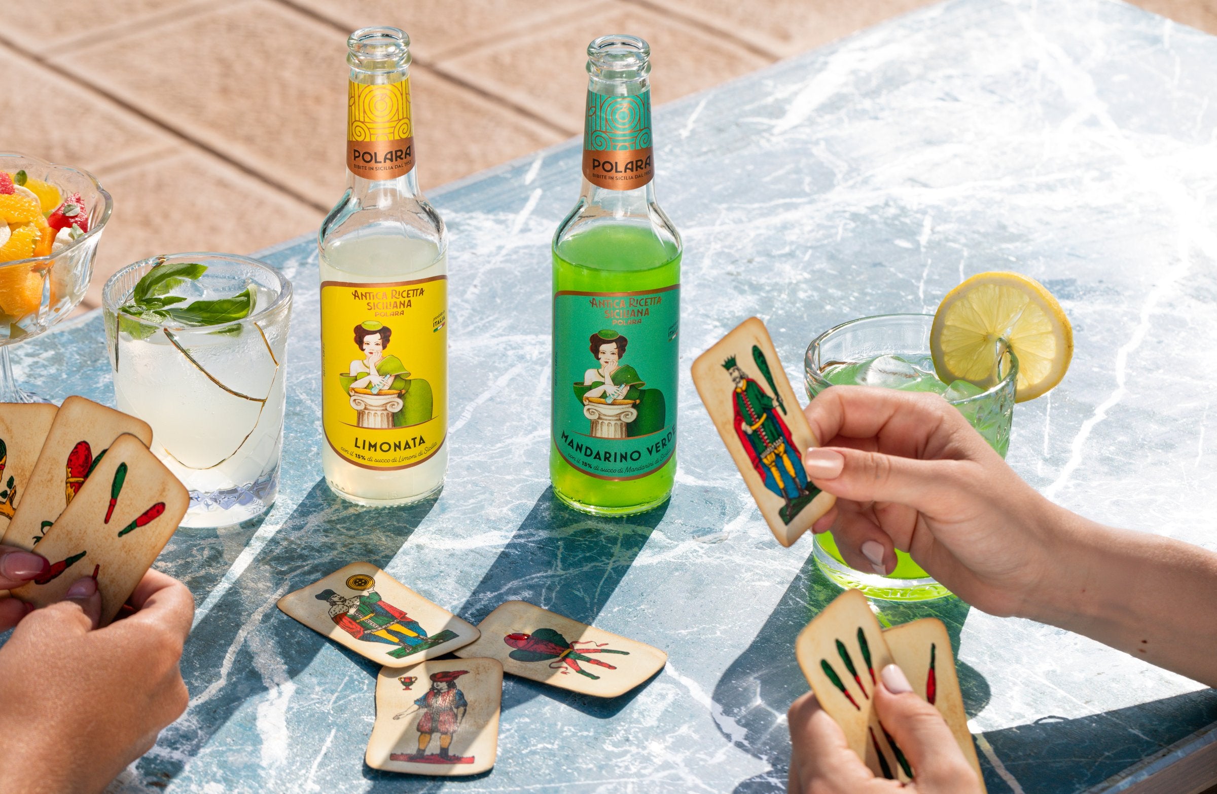 Bottles of Polara Limonata and Mandarino Verde on marble with playing cards and a lemon