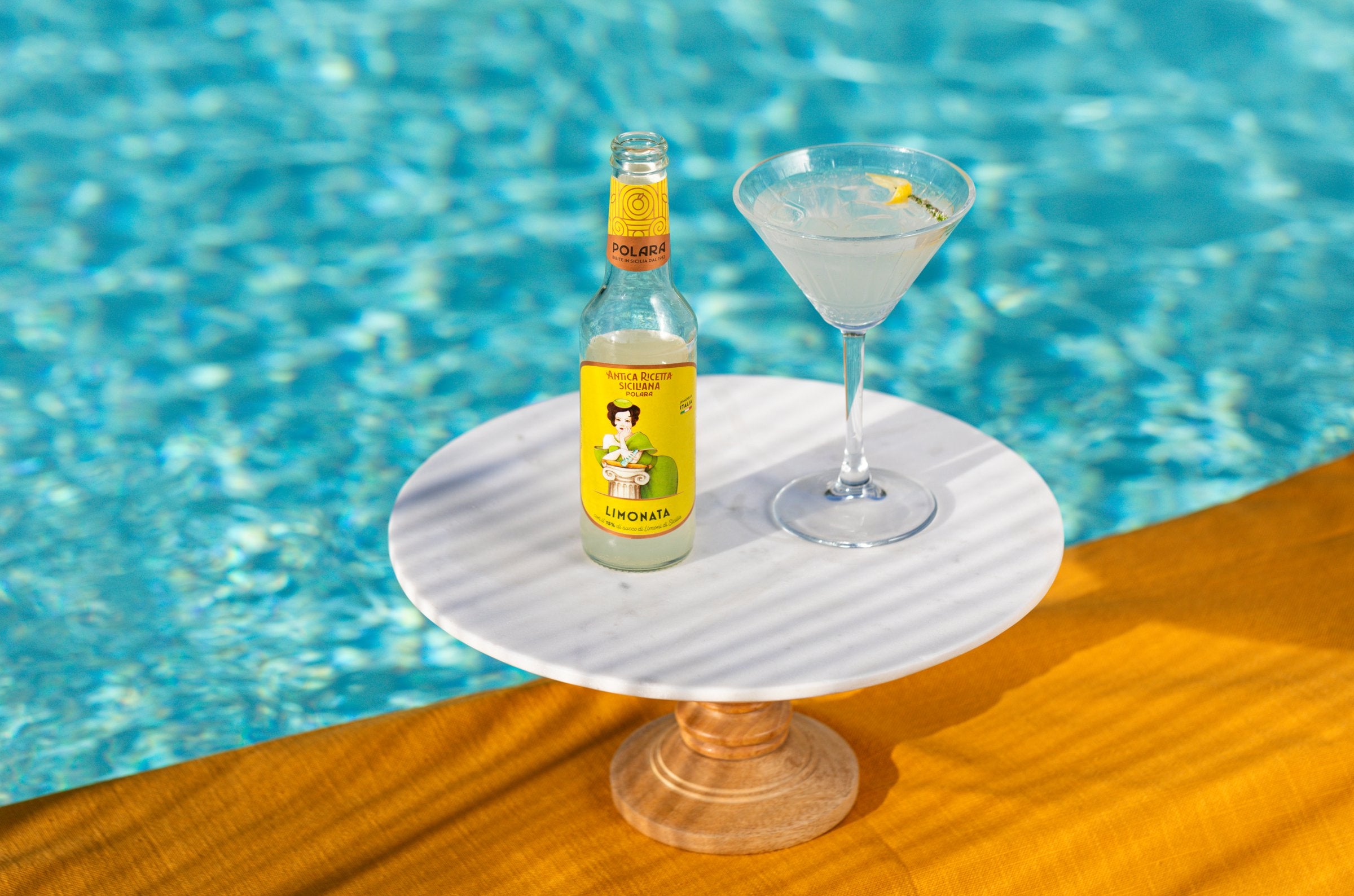 A bottle of Polara Limonata and a cocktail glass on a marble cake stand at the edge of a pool