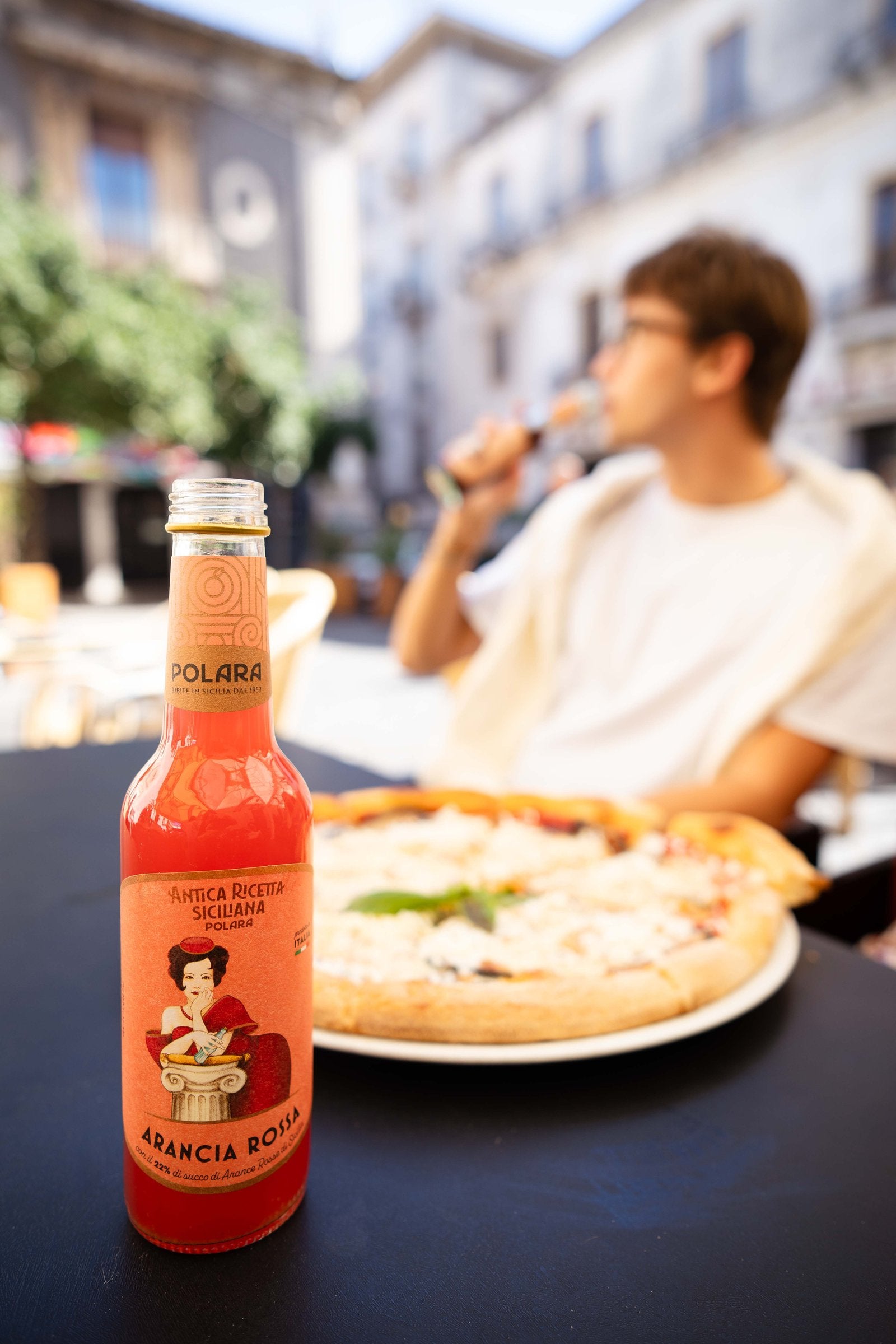 A bottle of Polara Aranciata Rossa beside a pizza on a café table in a town square, a young man drinking in the background