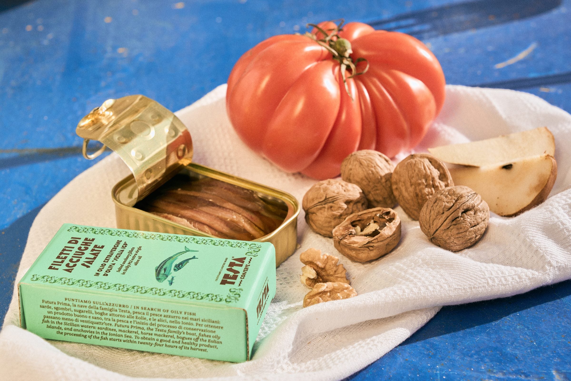 An open tin of Testa Ionian anchovy fillets beside a ripe tomato and walnuts on a linen cloth