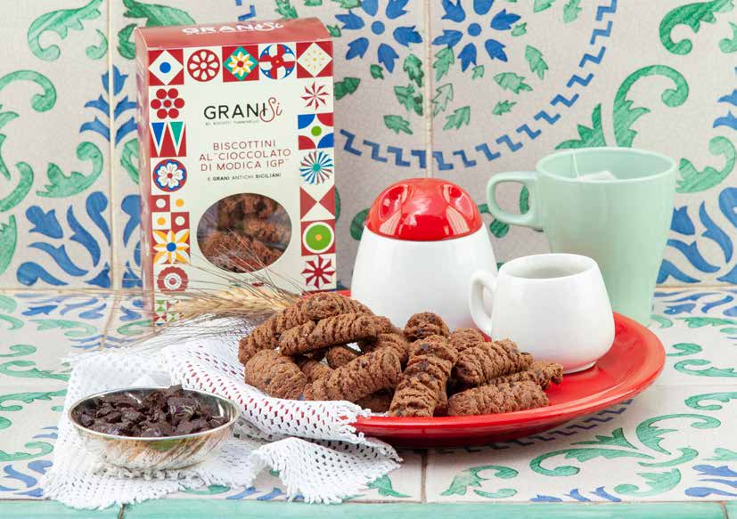 A box of Modica chocolate biscuits, the biscuits on a red plate beside a sugar bowl and a cup, on painted tiles